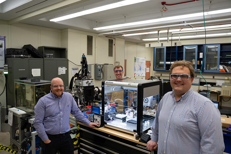 Three men in safety glasses pose standing around a large clear plastic box on a lab table. The box holds several small pieces of equipment. 