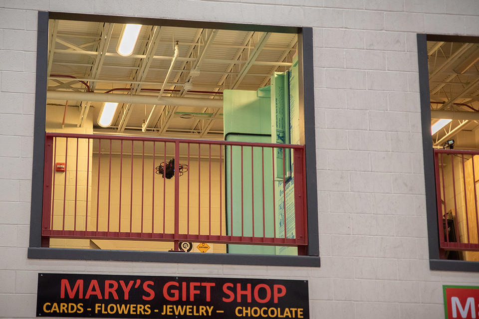 A drone is flying inside a second-story balcony space in a testing facility that includes pretend storefronts like Mary's Gift Shop. 