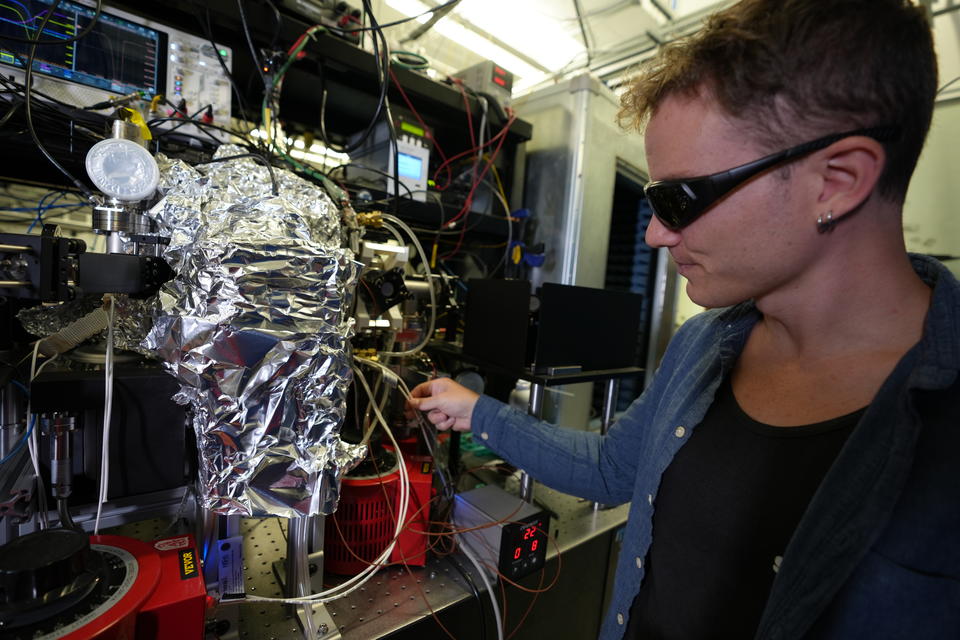 Noah Schlossberger wears dark safety glasses as he stands in front of a complicated electronic device in the lab. The device has wires coming out and is partially wrapped in aluminum foil. 