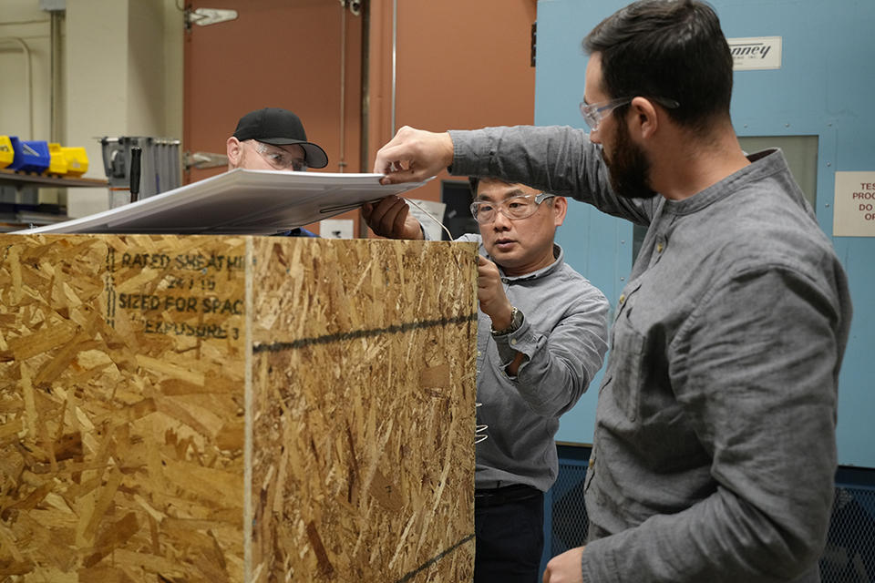 Three men wearing safety glasses stand around a wooden box they are assembling in the lab.