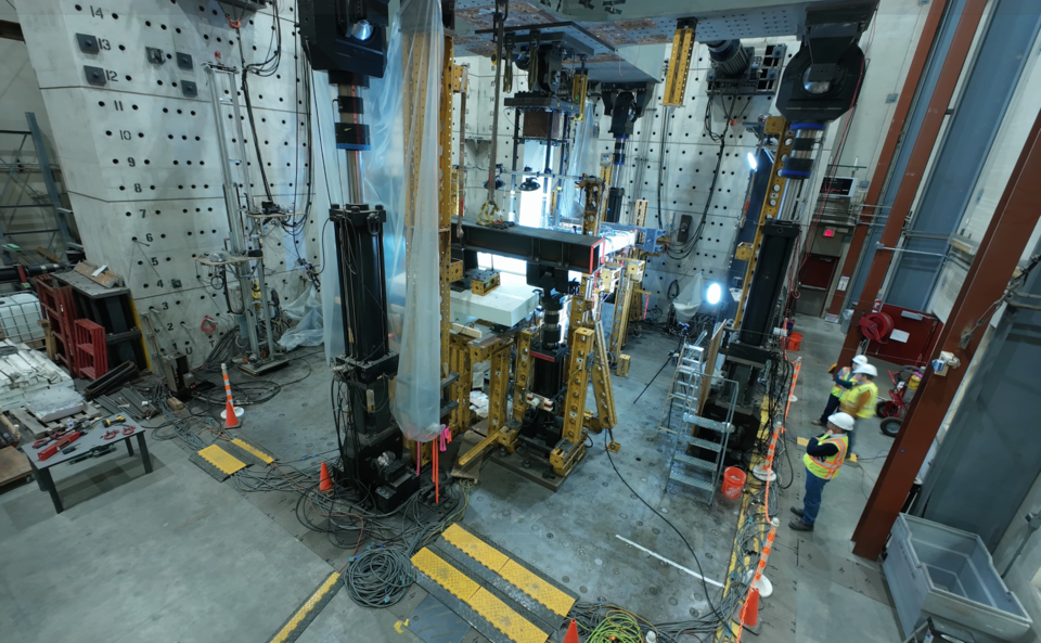 A high-ceilinged lab space is filled with large pieces of equipment, including a square structure in the center. A few people in hard hats stand at the edge of the room. 