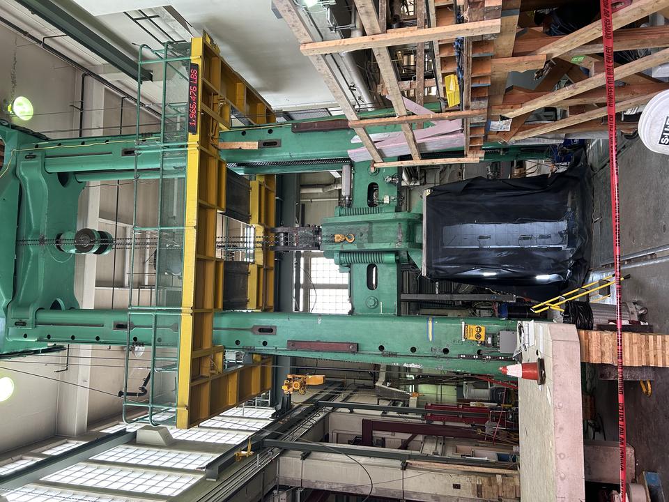 A two-story metal testing apparatus stands inside a high-ceilinged lab space filled with other equipment and scaffolding. 