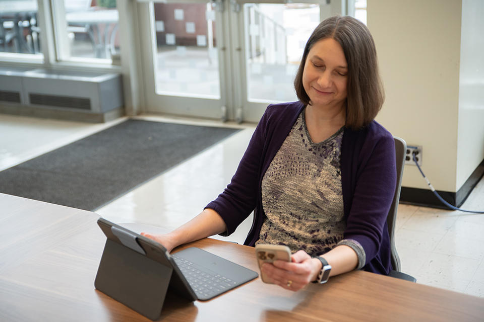 Julie Haney sits at a table in the NIST library with a laptop, looking at a smartphone in her hand. 