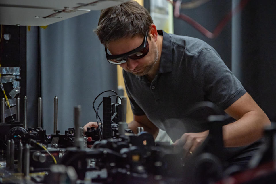 Krister Shalm wears safety glasses as he leans over a lab table covered with electronics equipment.