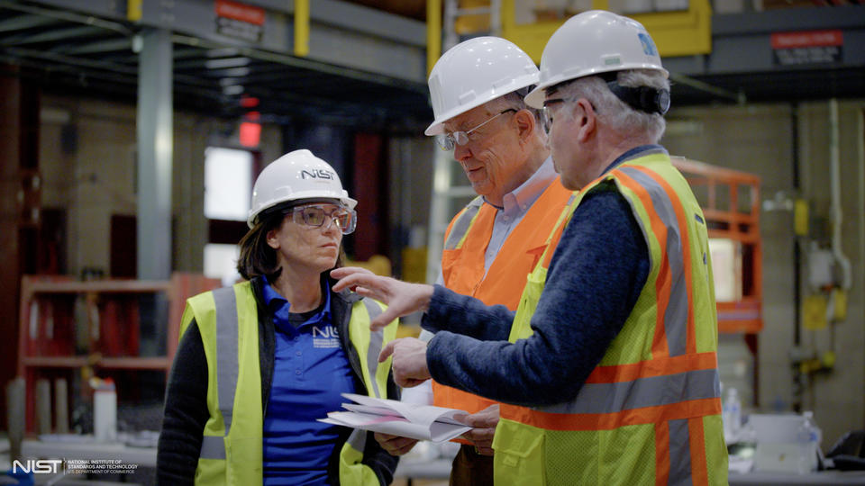 Three team members in construction safety gear stand talking inside the large warehouse where materials testing is taking place.