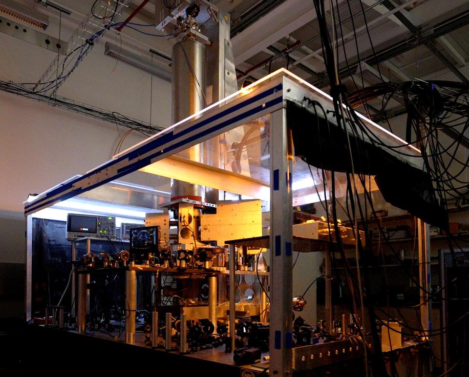 A large clear-sided box inside a darkened lab holds electronic equipment, wiring, and a metal cylinder rising up from the box. 