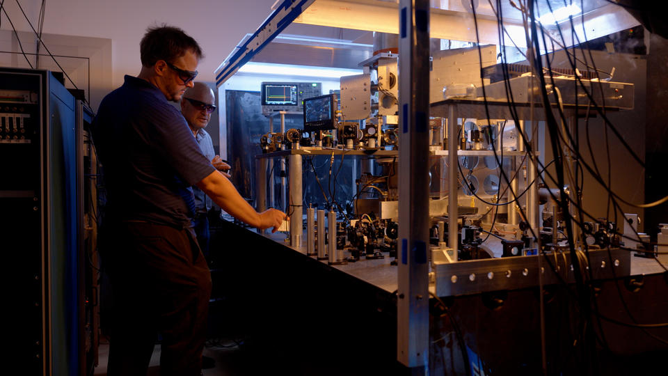 Two researchers in safety glasses stand at the edge of a metal table holding electronic and laser devices, wiring and metal structures.