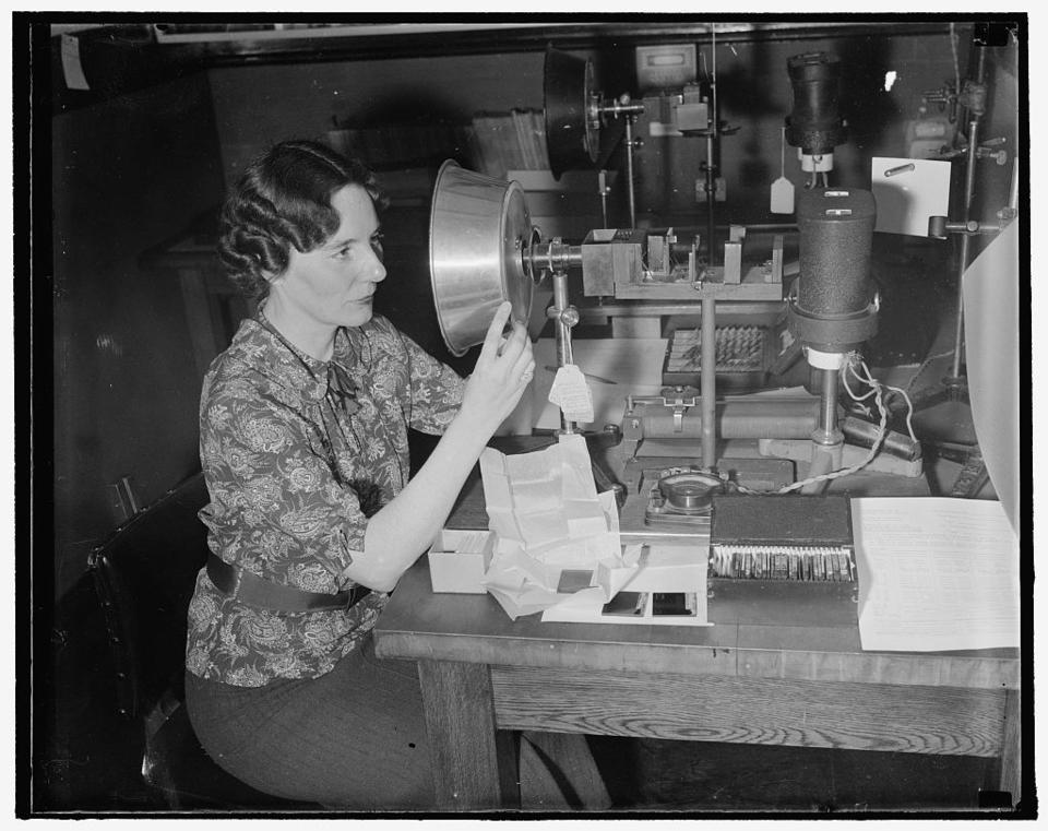 Historical photo shows woman adjusting a large metal lamp at a table full of similar equipment. 