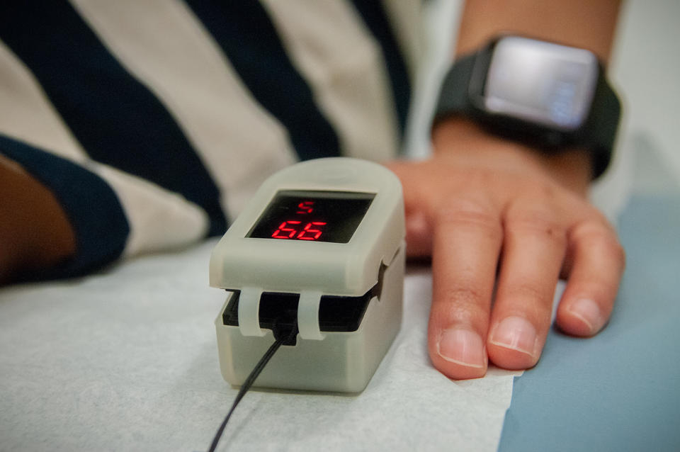 Close-up of a clip-on medical device on the finger of a person sitting on an exam table. The device screen reads 99. 
