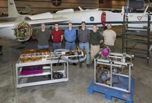 Six men stand in front of an airplane in a hangar, with equipment displayed on carts in front of them. 