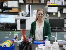 Briana Capistran wears safety glasses as she poses smiling in the lab with computers and other equipment in the background. 