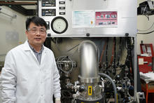 A researcher in a lab coat and safety glasses poses next to a large piece of lab equipment.