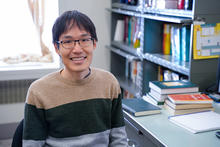 Leroy Jia poses smiling at his desk, with piles of books in the background. 