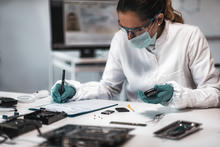 A researcher wearing a mask, safety glasses and gloves sits at a desk, looking at electronic devices and writing on a clipboard. 