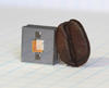 Square gray cube with two square holes in it and a gold square in between the holes, and a dark brown coffee bean leaning against the cube.