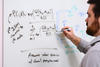 Man with dark hair writing mathematical formulas on a whiteboard on a wall.