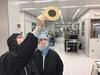 Two people wearing coveralls in a "clean room" lab look up at a disk-shaped object that one of them is holding. 