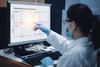 A researcher wearing a mask and gloves points at a computer screen showing graphs labeled "Mass Spectrometry Analysis Report."