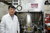 A researcher in a lab coat and safety glasses poses next to a large piece of lab equipment.